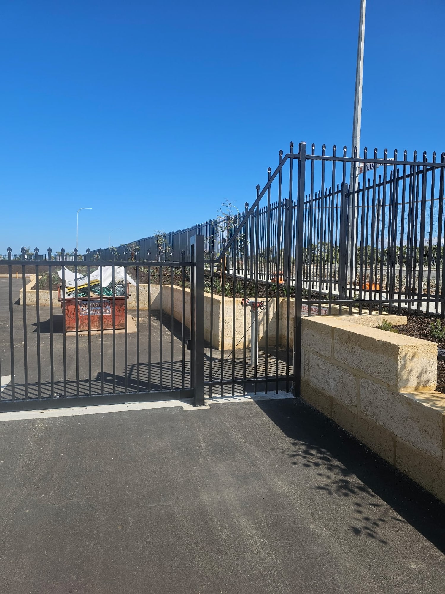 Black garrison security fence and swing gate above a limestone retaining wall at a commercial carpark