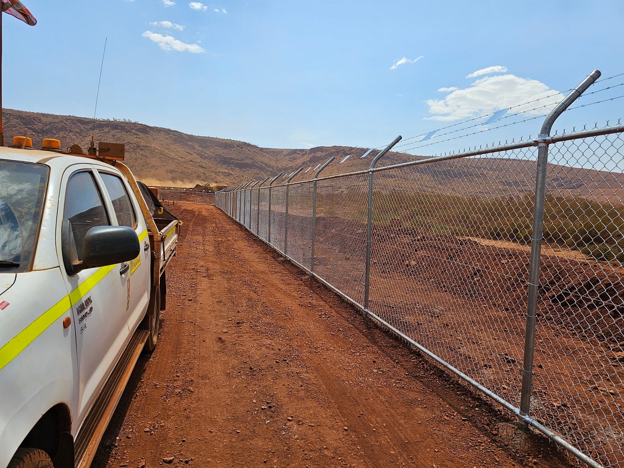 Long galvanised chainmesh fence with barbed top arms running across a Pilbara mine site