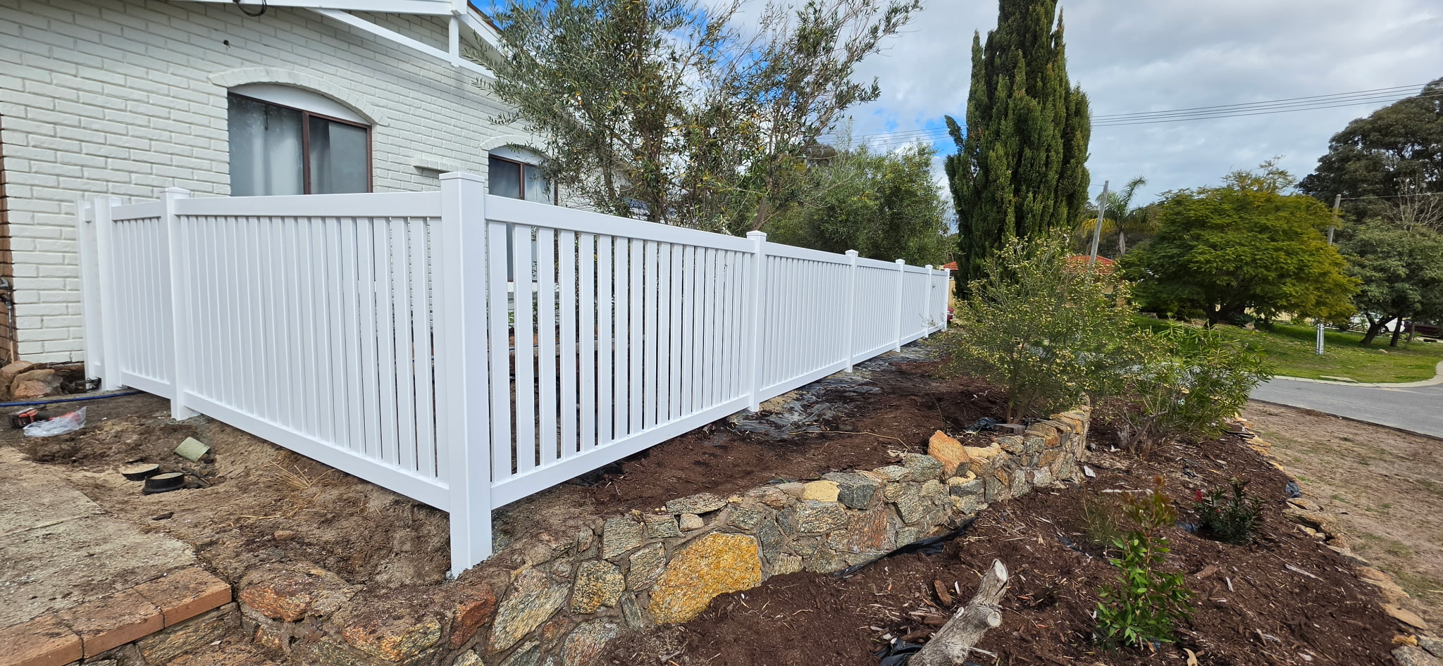 White PVC picket fence turning the corner of a front yard with new garden beds and a stone retaining wall